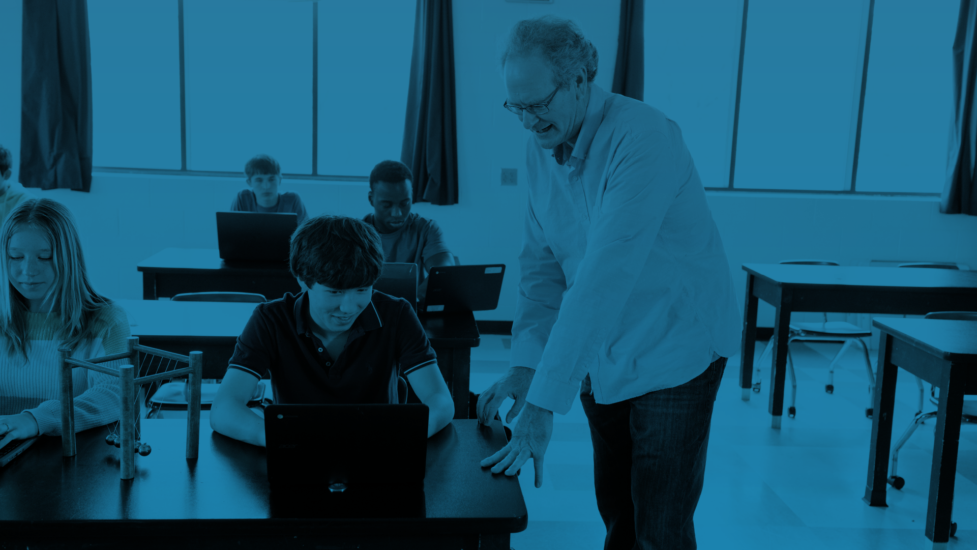 A professor standing next to a student using a laptop