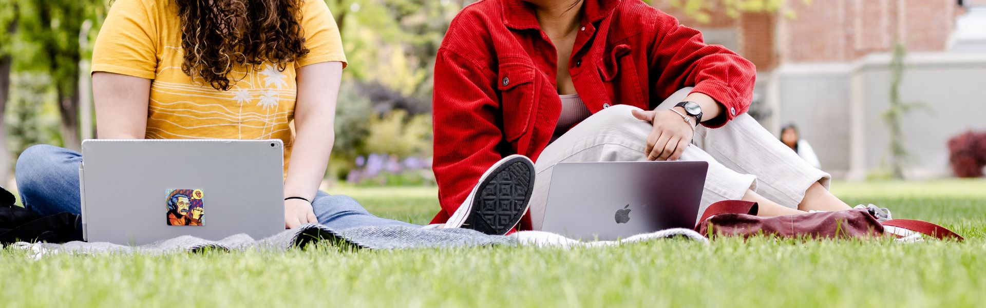 image of two girls, one in a yellow shirt and the other in a red jacket, sitting on the grass with their laptops open in front of them.