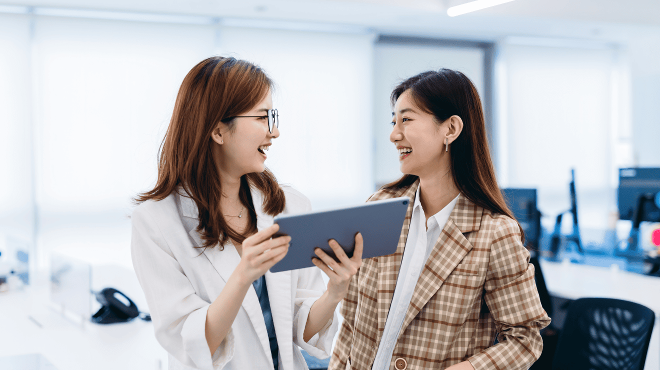 Two women collaborating in an office, talking while using a tablet.