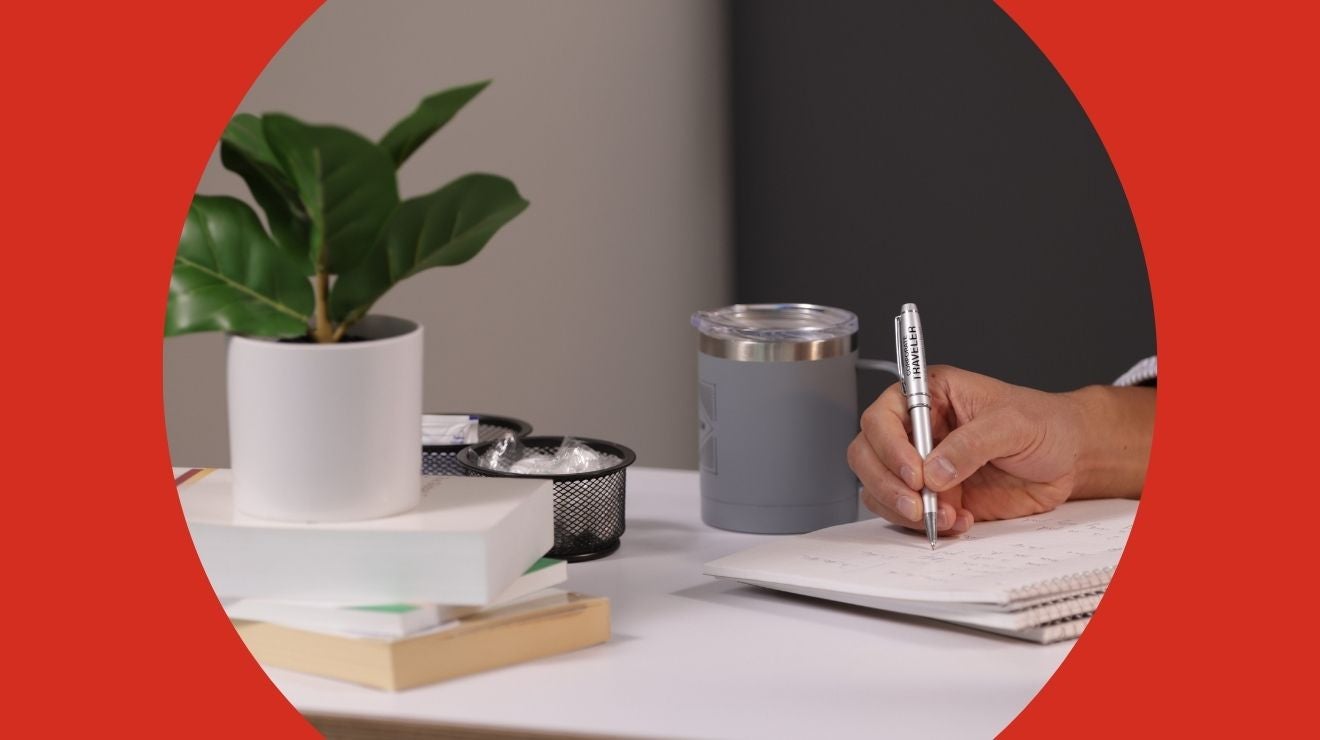 Hand writing in notebook next to a plant and coffee mug.