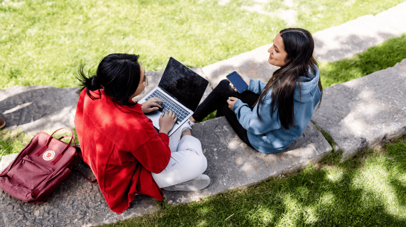 two students talking while using a laptop and a cellphone