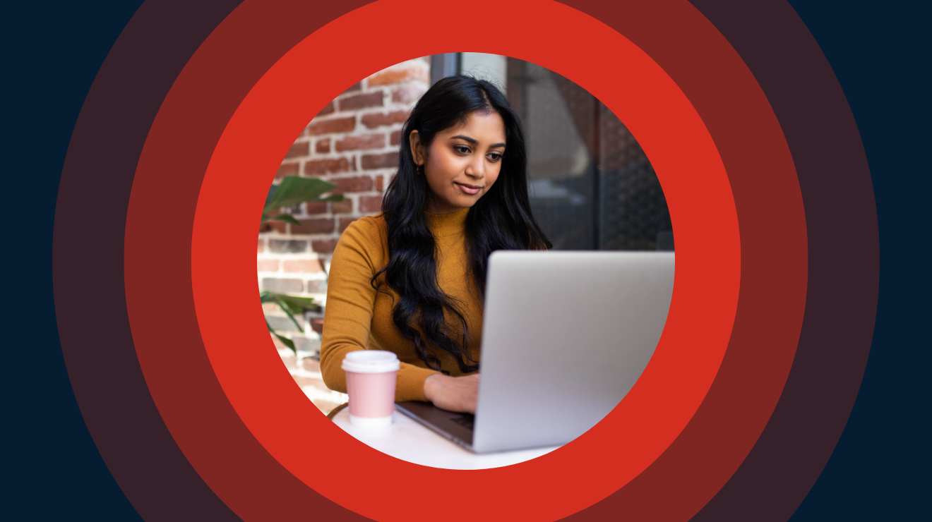 A woman sitting at a table, focused on her laptop, with a calm expression in a well-lit room.