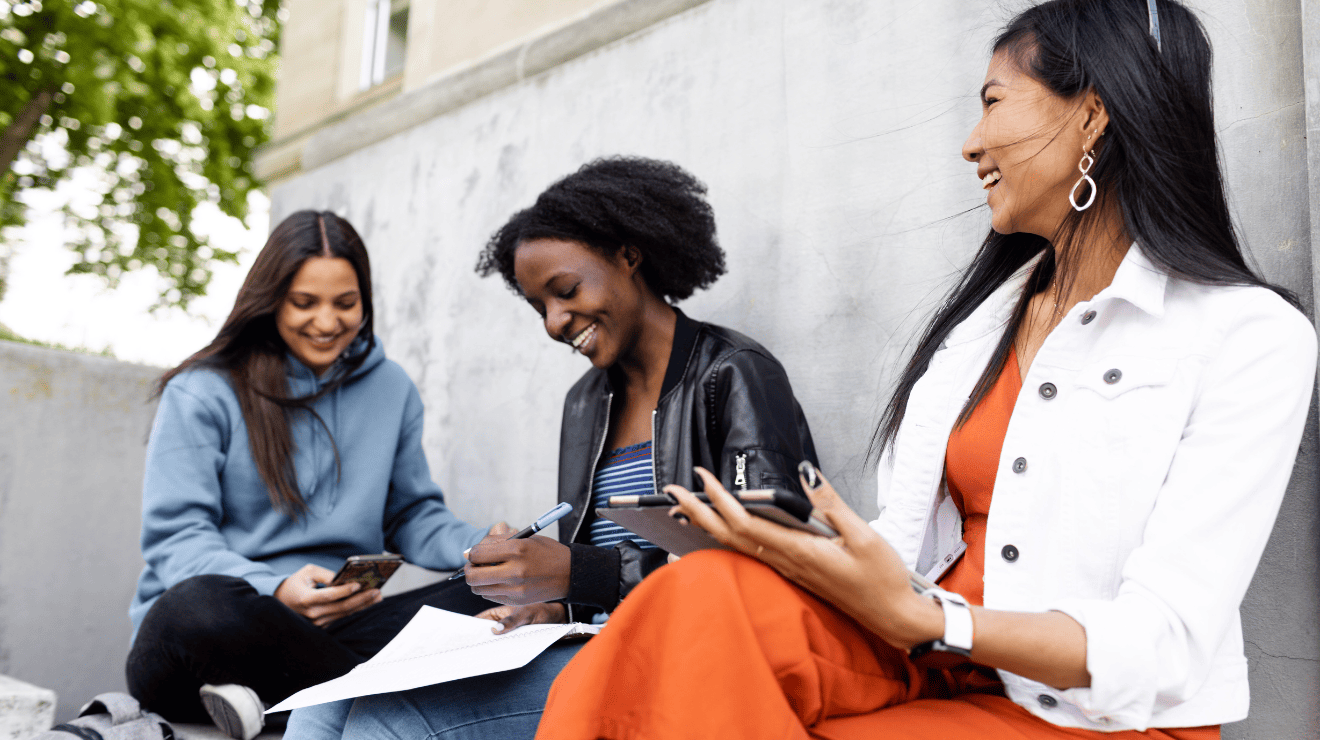 Three young women sit on a wall, each focused on their phones, enjoying a moment together outdoors.
