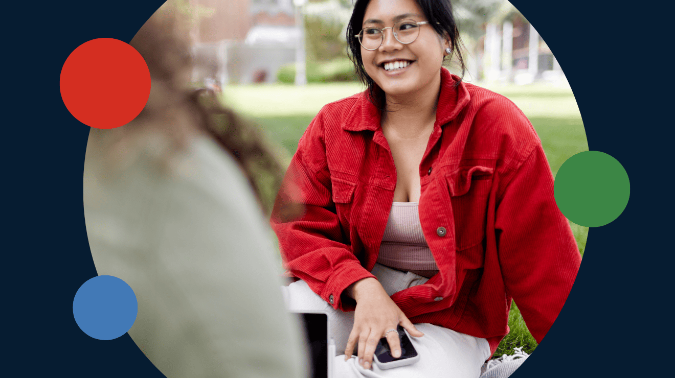 A woman sitting on the grass, holding a phone, appears relaxed and engaged in her surroundings.