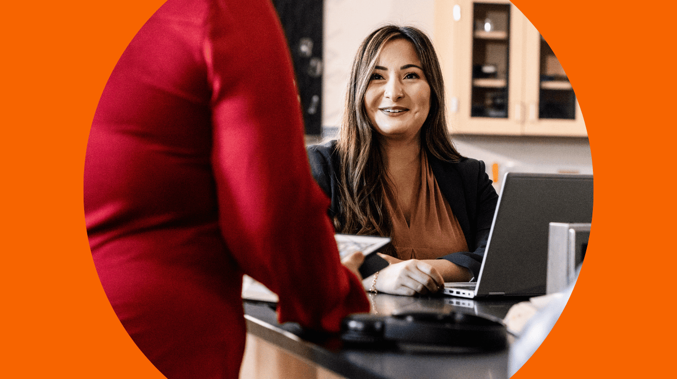 A woman talks to another woman at a desk, indicating a professional or collaborative interaction.