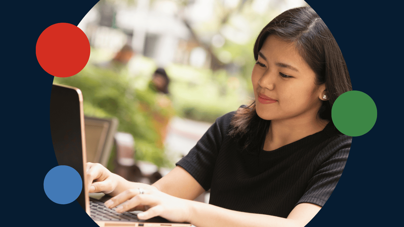 A woman seated at a table, focused on her laptop, with a calm expression in a well-lit environment.