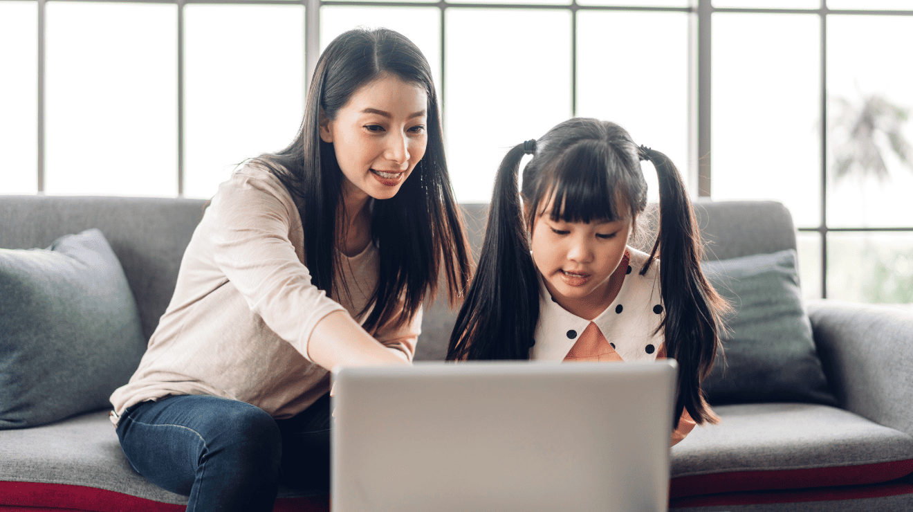 A woman and a young girl sit together on a couch, focused on a laptop screen in front of them.