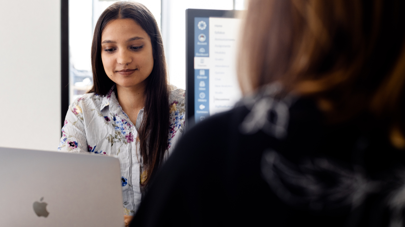 A woman seated at a desk, focused on her laptop, with a notepad and pen beside her.