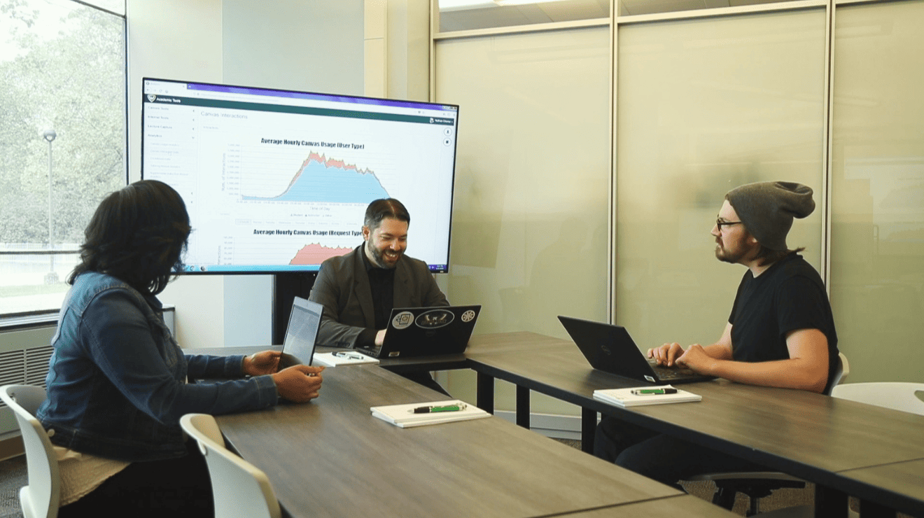 Three individuals seated at a table, each using a laptop, engaged in discussion or collaboration.