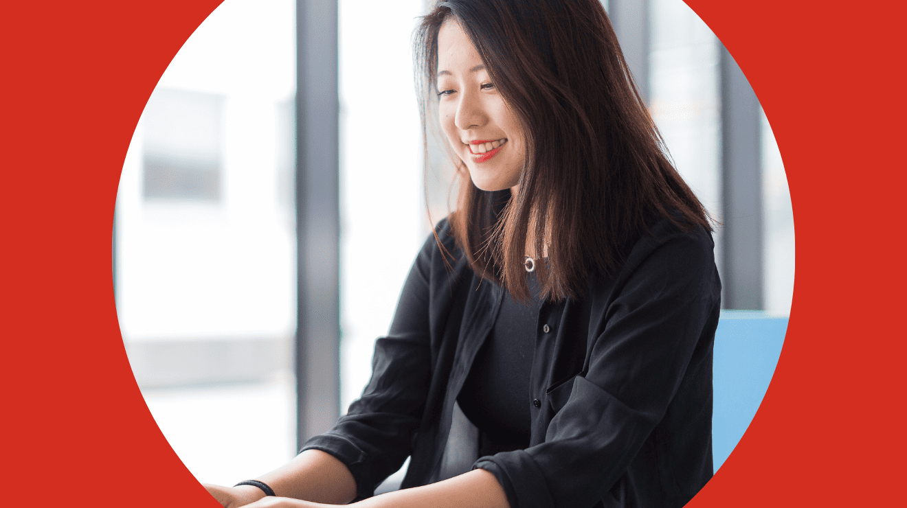 A woman seated at a desk, focused on her laptop, with a notepad and pen beside her.