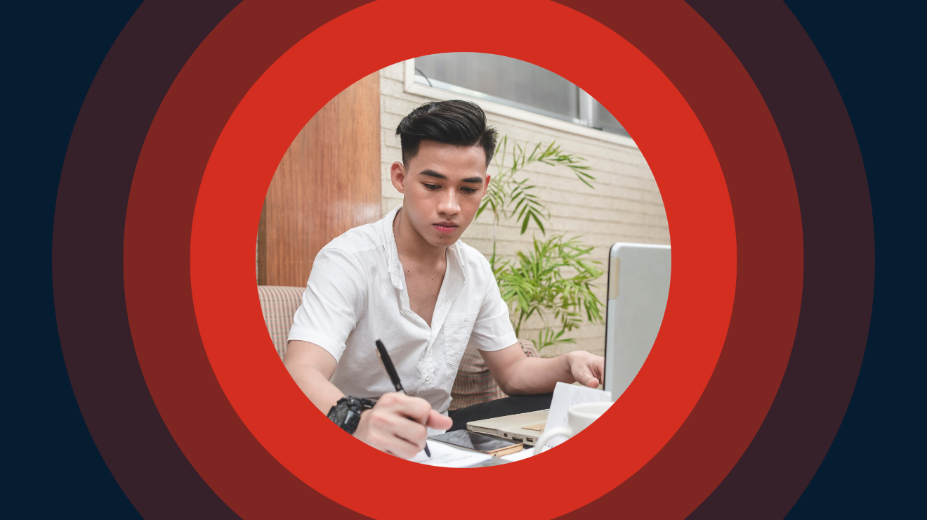 A man seated at a desk, working on a laptop with a pen in hand, focused on his task.