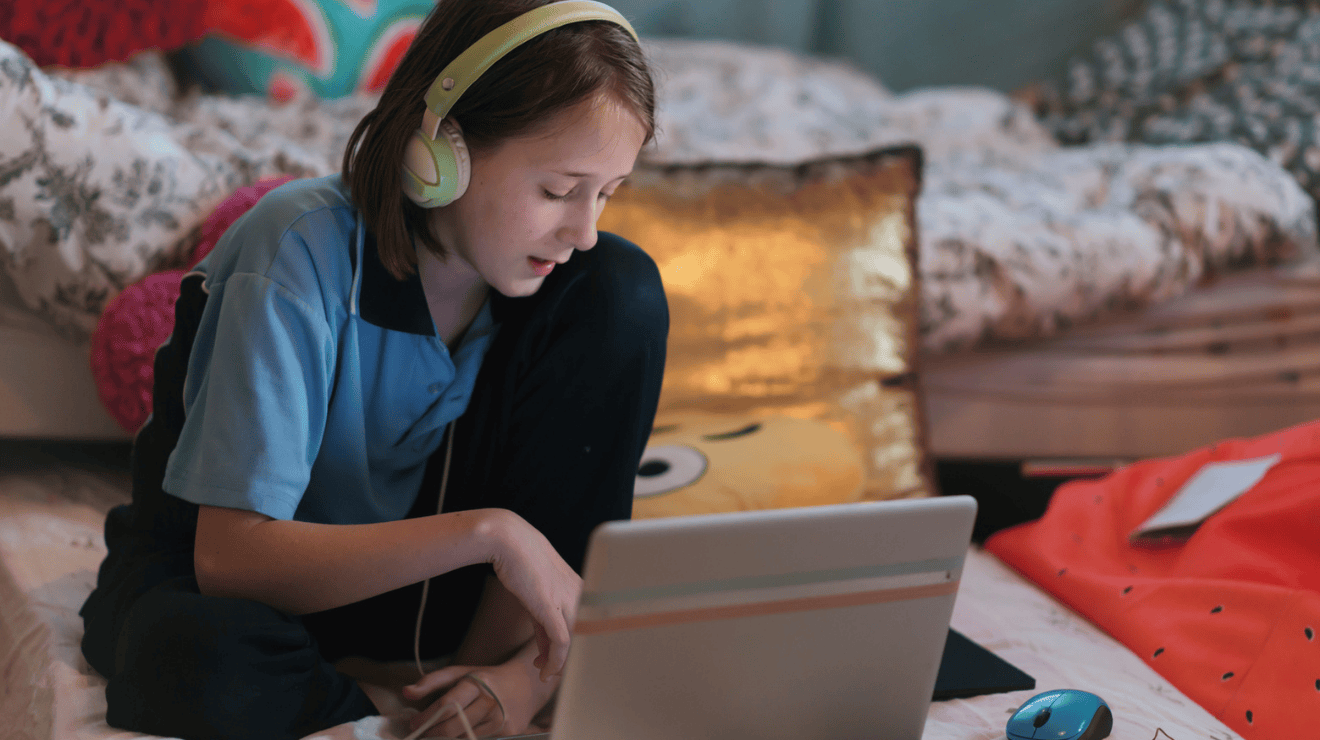 School-aged learner using a laptop at home, wearing headphones and focusing on an online lesson while seated on a bed.