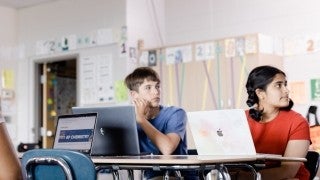 Two students sitting at a desk, laptops out, looking at teacher during a lesson