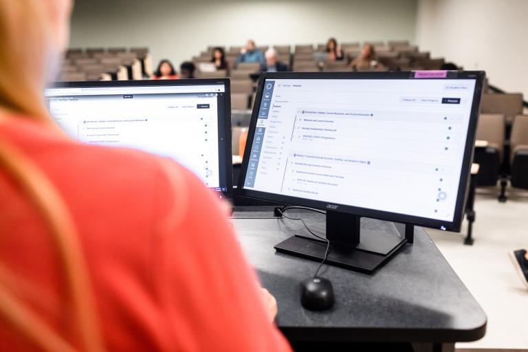 A professor looks at their computer while standing in front of the classroom.