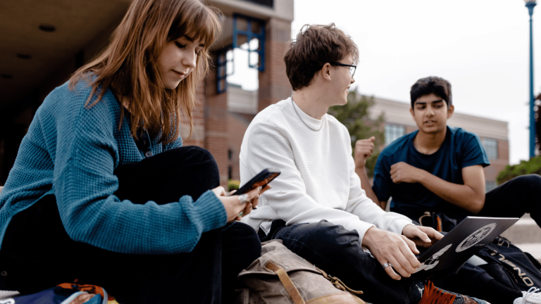 three students sitting on steps, talking to each other and looking at laptops and phones