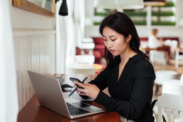 Woman in coffee shop reads her phone while looking at a laptop