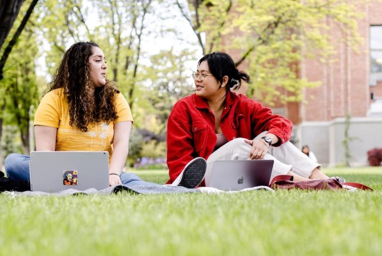 two students sit with laptops in the grass together