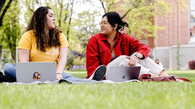 two students sit with laptops in the grass together