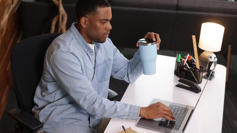 man in a blue long sleeve shirt holding his cup while typing on his computer at his desk