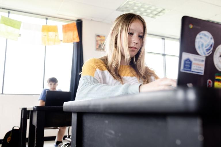 Students sit at desks, taking an assessment on laptops.