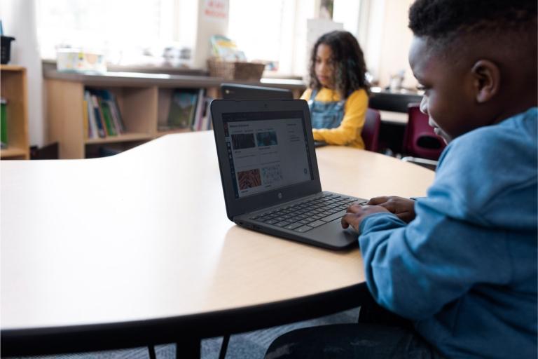 Two students sit at a table using laptops.