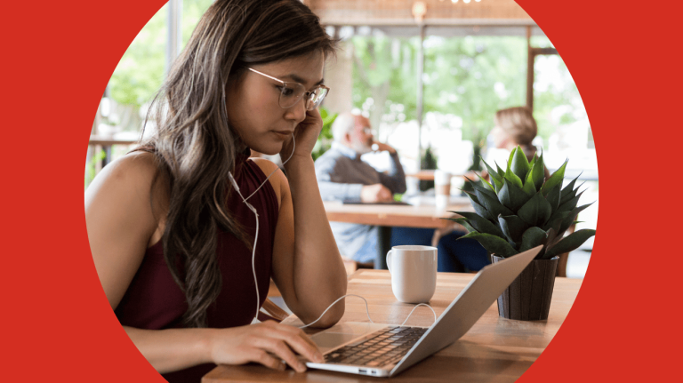 A woman in a red shirt sits at a table, working on a laptop with a focused expression.