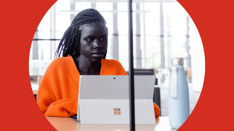 A woman in an orange shirt sits at a desk, working on a laptop.