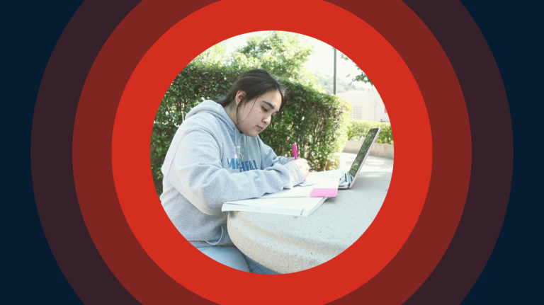 A woman seated at a table, focused on her laptop, with a calm expression in an outdoor setting.