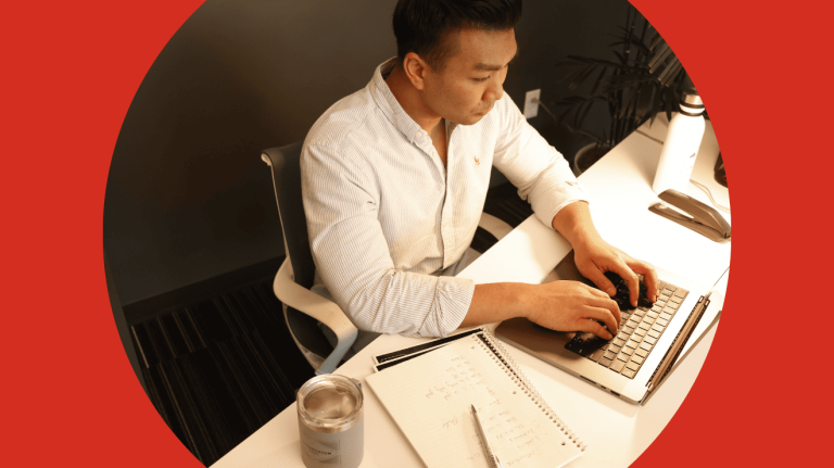 A man sitting at a desk, focused on his laptop, with a notepad and pen beside him.