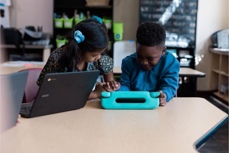 Two students sit at a table using a tablet