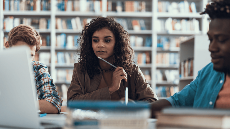 Three students collaborate on a laptop while seated at a table in a library, surrounded by bookshelves.