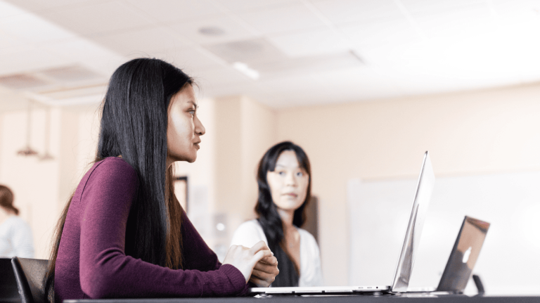 Two women engaged in conversation while working on laptops at a table.