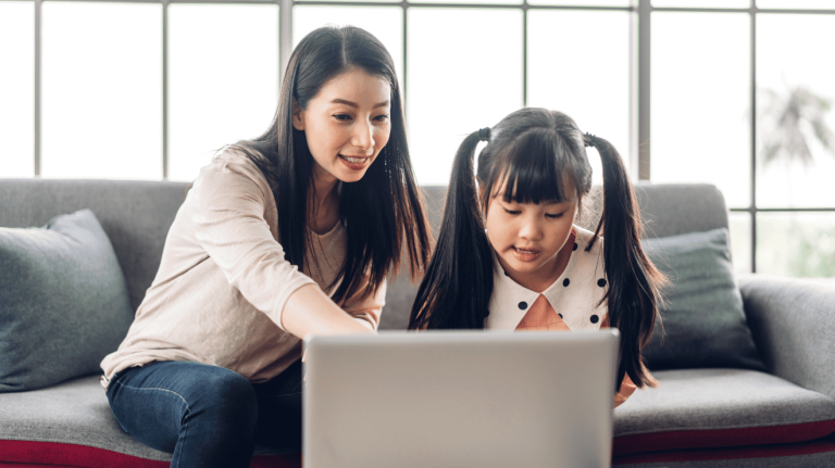 A woman and a young girl sit together on a couch, focused on a laptop screen in front of them.