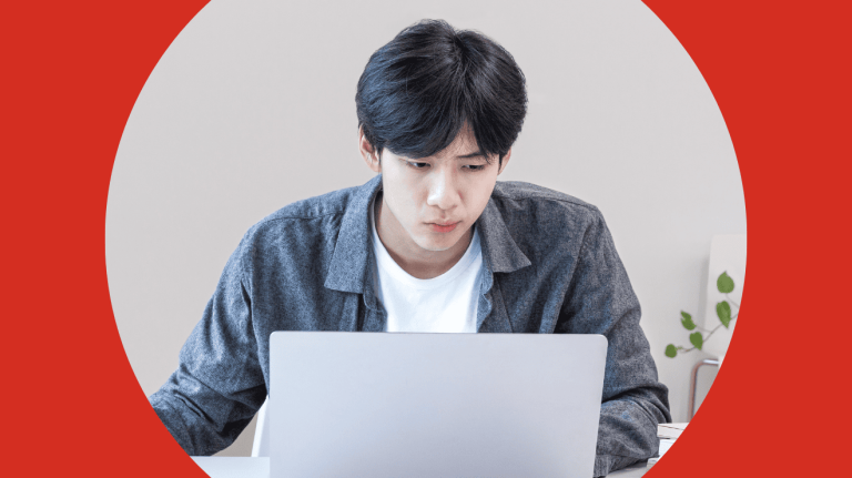 A man seated at a desk, focused on his laptop, with a notepad and pen beside him.