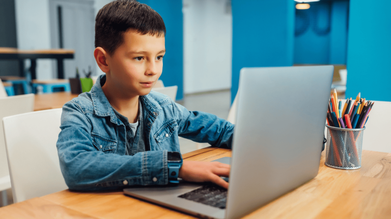 A young boy sitting at a table, focused on his laptop, with a thoughtful expression on his face.