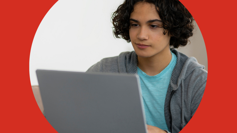 A young man with curly hair is engaged with a laptop, looking thoughtfully at the screen in a study environment.
