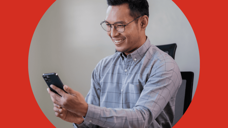 A smiling man in glasses is using his phone, appearing engaged and happy in a casual setting.