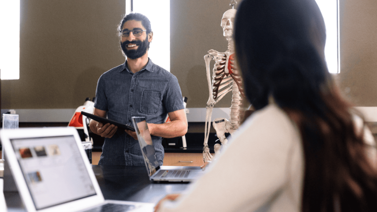 A man and woman are engaged with a computer, with a skeleton positioned on the desk nearby.