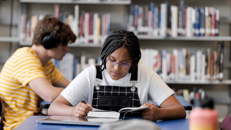 A young girl sits in a library, engrossed in a book, surrounded by shelves filled with various books.