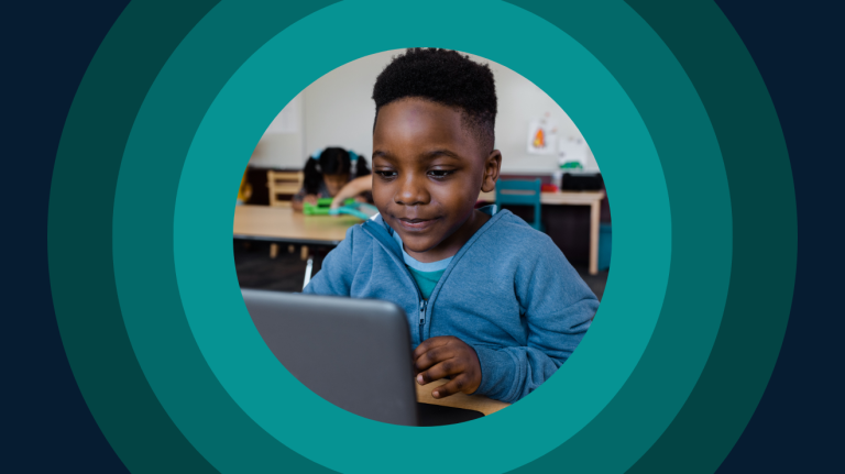 A young boy focused on a laptop while seated at a desk in a classroom setting.