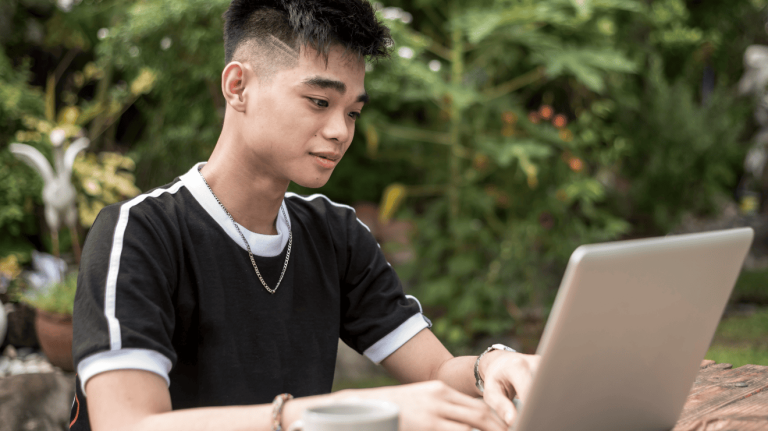 A young man seated at a table, focused on his laptop while working or studying.