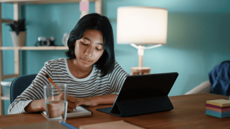 A young woman sits at a table, focused on her laptop, with a thoughtful expression on her face.