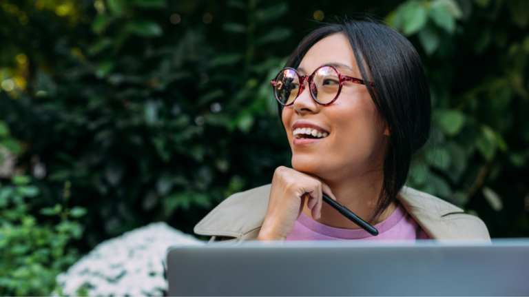 A woman with glasses smiles while working on her laptop, conveying a sense of joy and engagement.