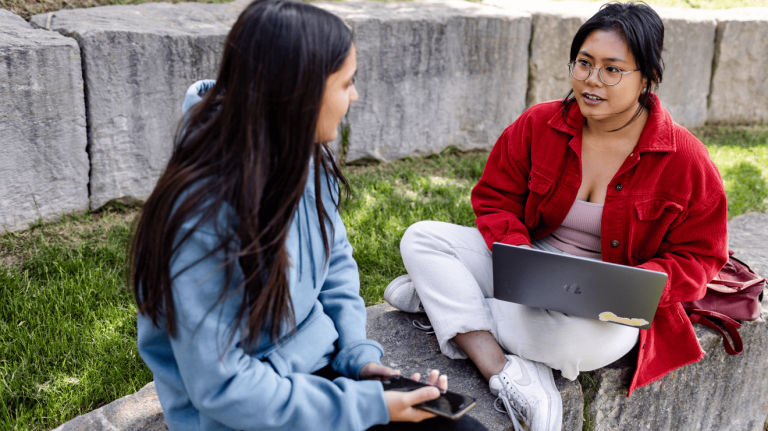 Two women sit on a stone wall, working together on a laptop in an outdoor setting.