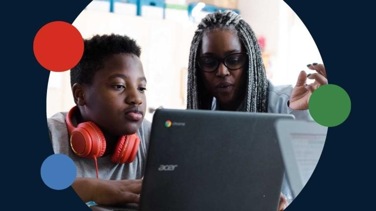 a teacher sits with a student at a laptop
