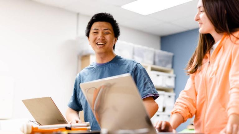 Two students sit at a table using laptops.