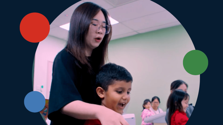A photo of Yamaha educator Shi Bei in a music classroom in Malaysia looking over the shoulder of a young student. 