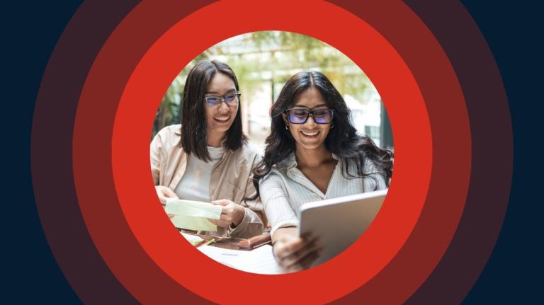Two smiling women in glasses collaborating and looking at a tablet. The photo is centered inside a graphic design of concentric circles in shades of red and dark blue.