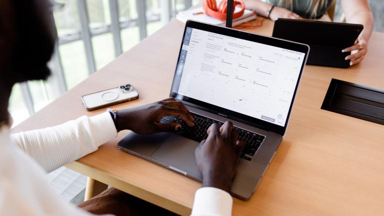 students sit at a table on a university campus at their laptops