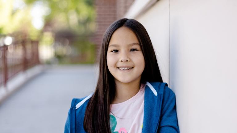 an elementary student stands outside of the school on the sidewalk and smiles at the camera